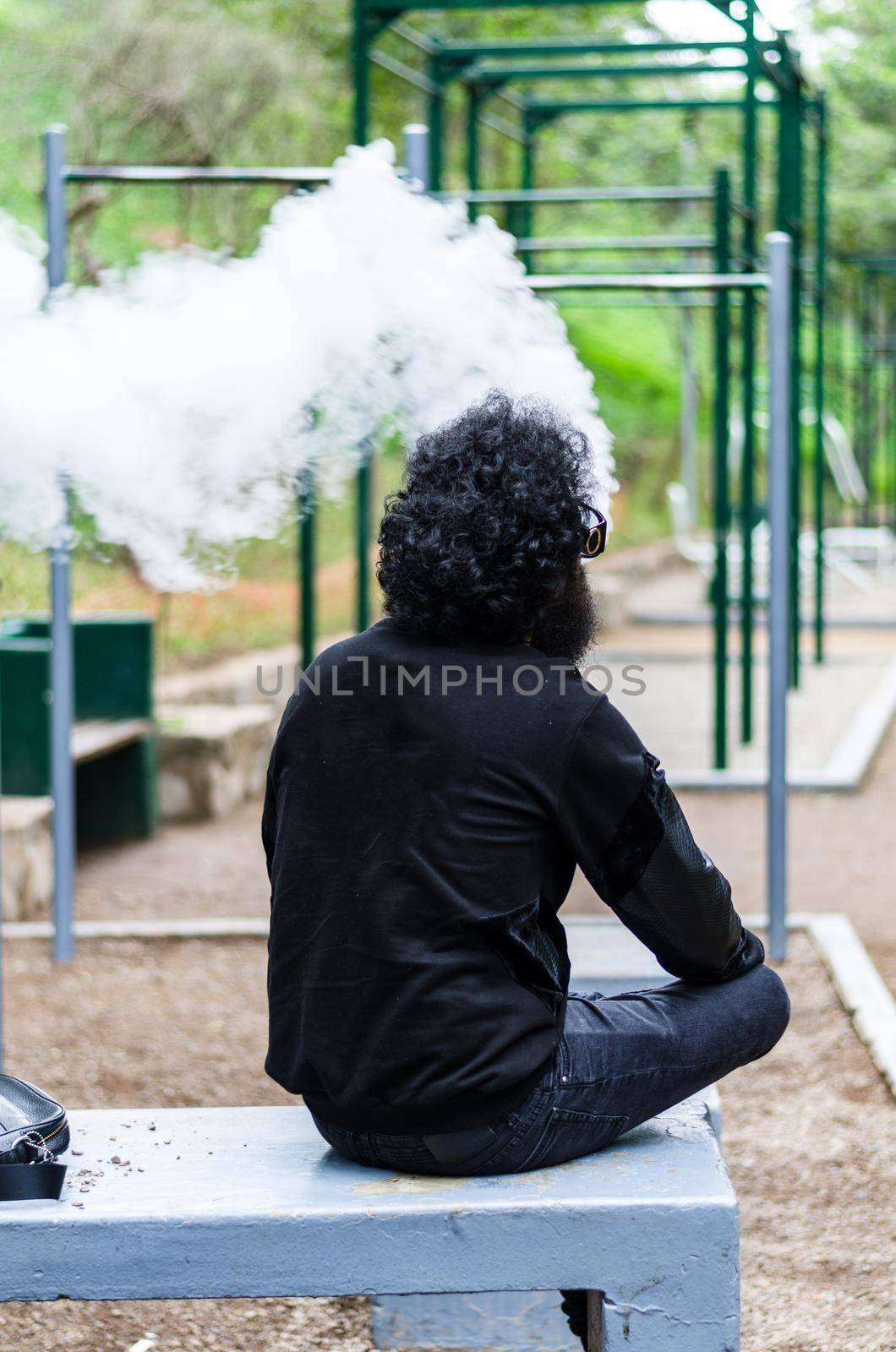 Man with a beard smokes an electronic cigarette sit on a bench in the park