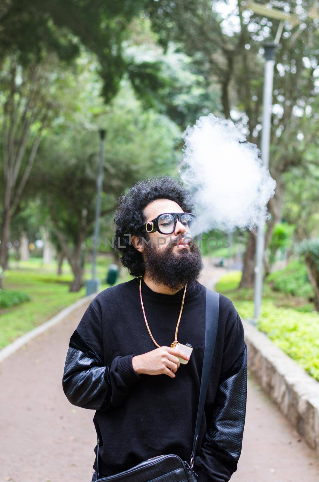 Vape. Young brutal man with large beard and fashionable haircut in sunglasses smoking an electronic cigarette in the city park. Steam cloud. Lifestyle.