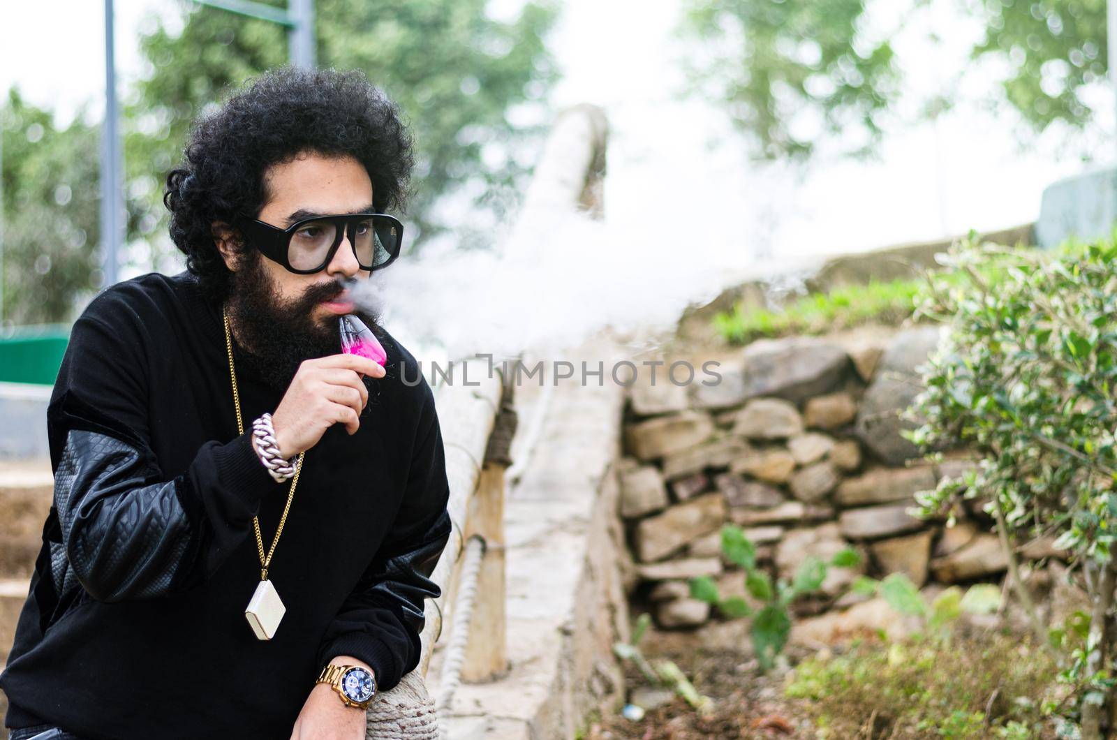 Vape man. Outdoor portrait of a young brutal white guy with large beard and in sunglasses letting puffs out of steam from an electronic cigarette opposite the old wooden fence in the village. Vaping.