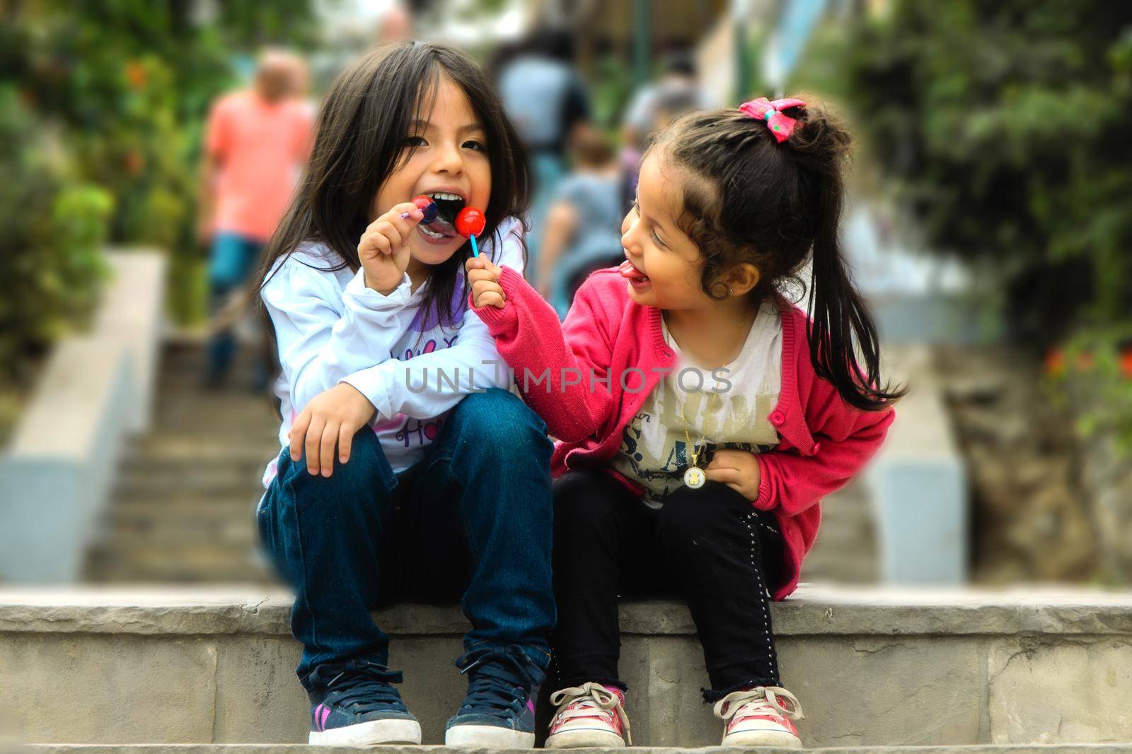 Two girls sitting and eating a lollipop in the park, summer outdoor portrait. best friends. by Peruphotoart