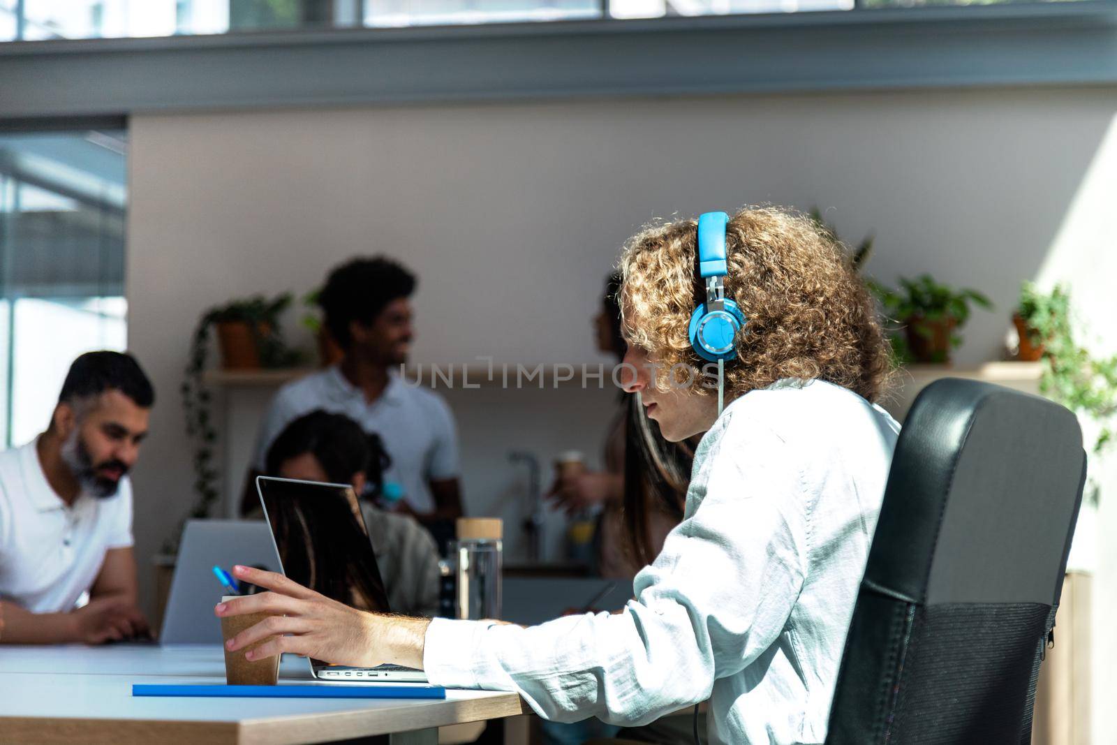 Young caucasian man with blue headphones working in busy office. Copy space. Startup concept. Coworking concept.