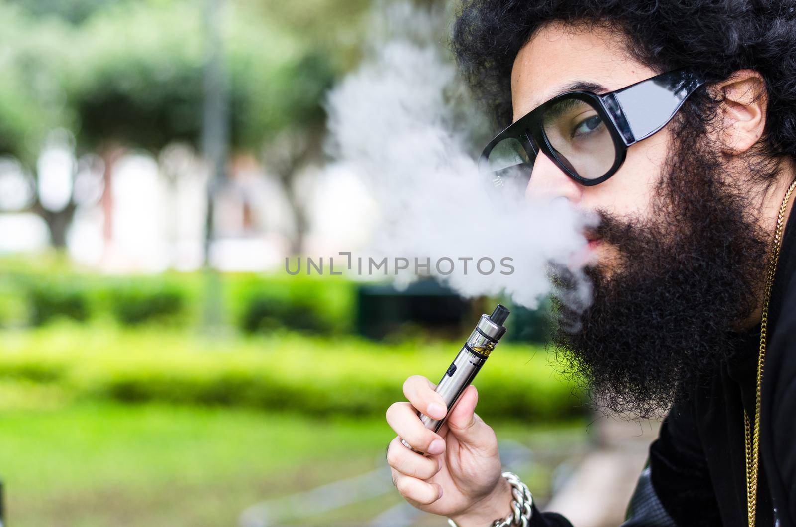 Bearded man smokes vape close up. Electronic cigarette concept. Man with long beard and clouds of smoke looks relaxed. Man with beard and mustache on calm face, branches on background, defocused.