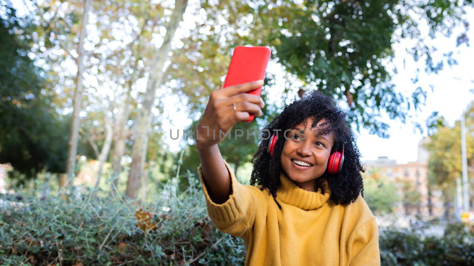 Panoramic image of young African American woman taking a selfie with smartphone in a park. Copy space. by Hoverstock