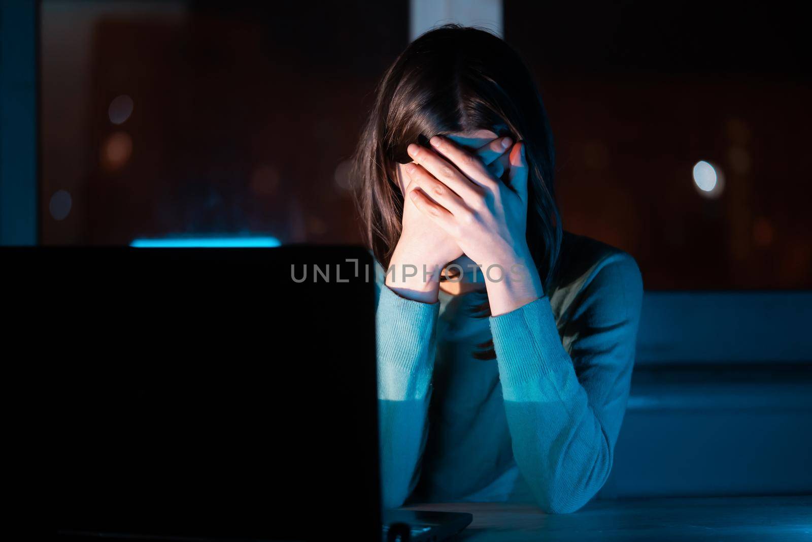 A young girl sits at a laptop at night at her home workplace in the dark and covers her face with her hands, the woman is upset, depressed, and has a headache, has some problems with a deadline.