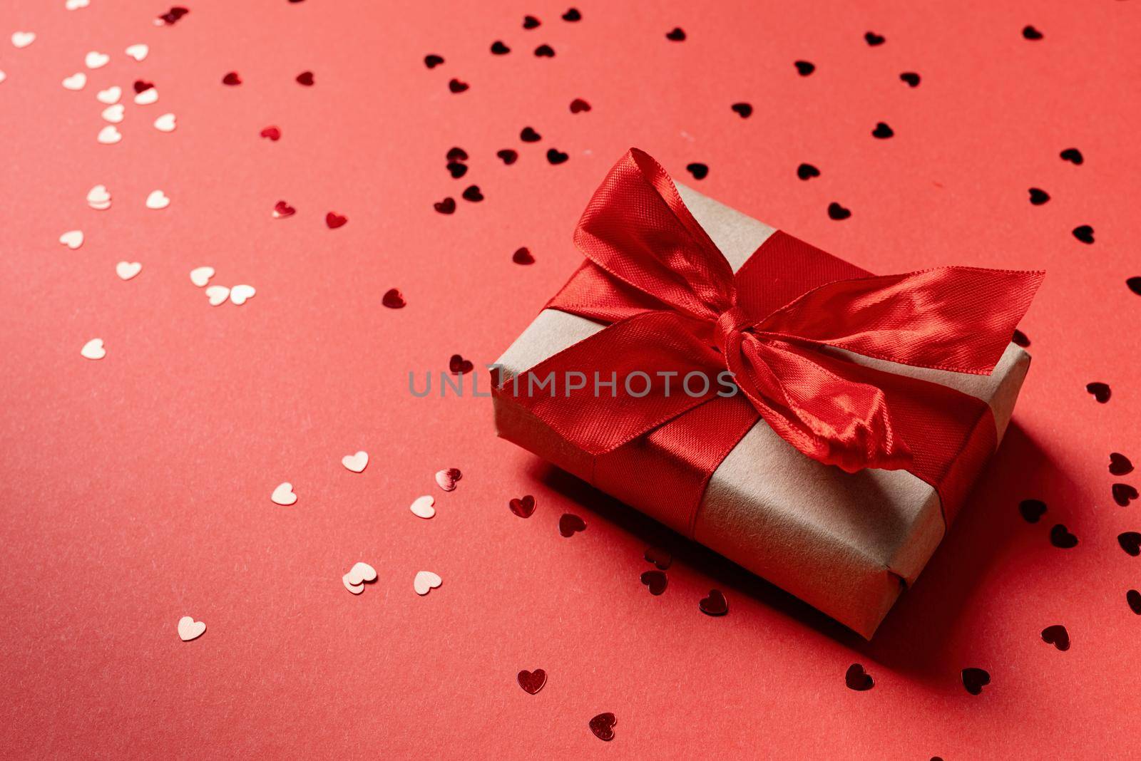 Valentines Day. Craft Gift box with beautiful red ribbon with heart shape confetti on red background with copy space