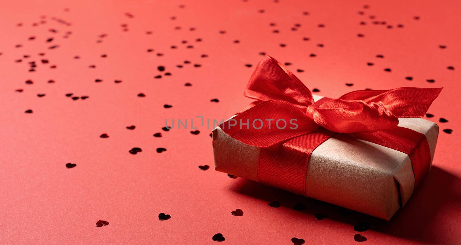 Valentines Day. Craft Gift box with beautiful red ribbon with heart shape confetti on red background with copy space