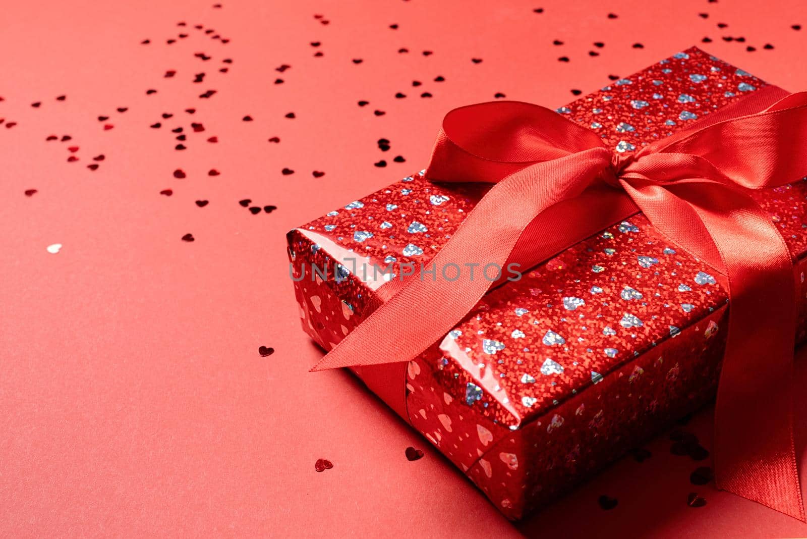 Valentines Day. Red Gift box with beautiful red ribbon with heart shape confetti on red background with copy space