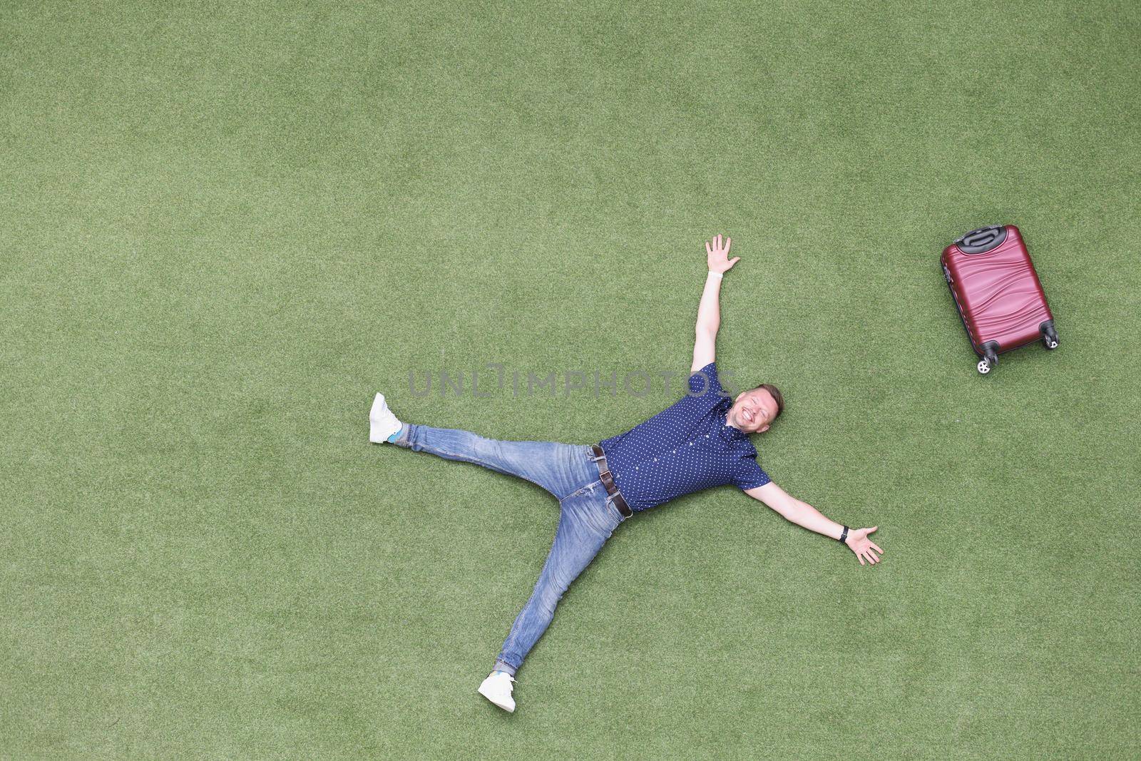 Happy man with a suitcase lies on a green lawn, top view. Arrival at the resort, vacation alone. Freedom from work