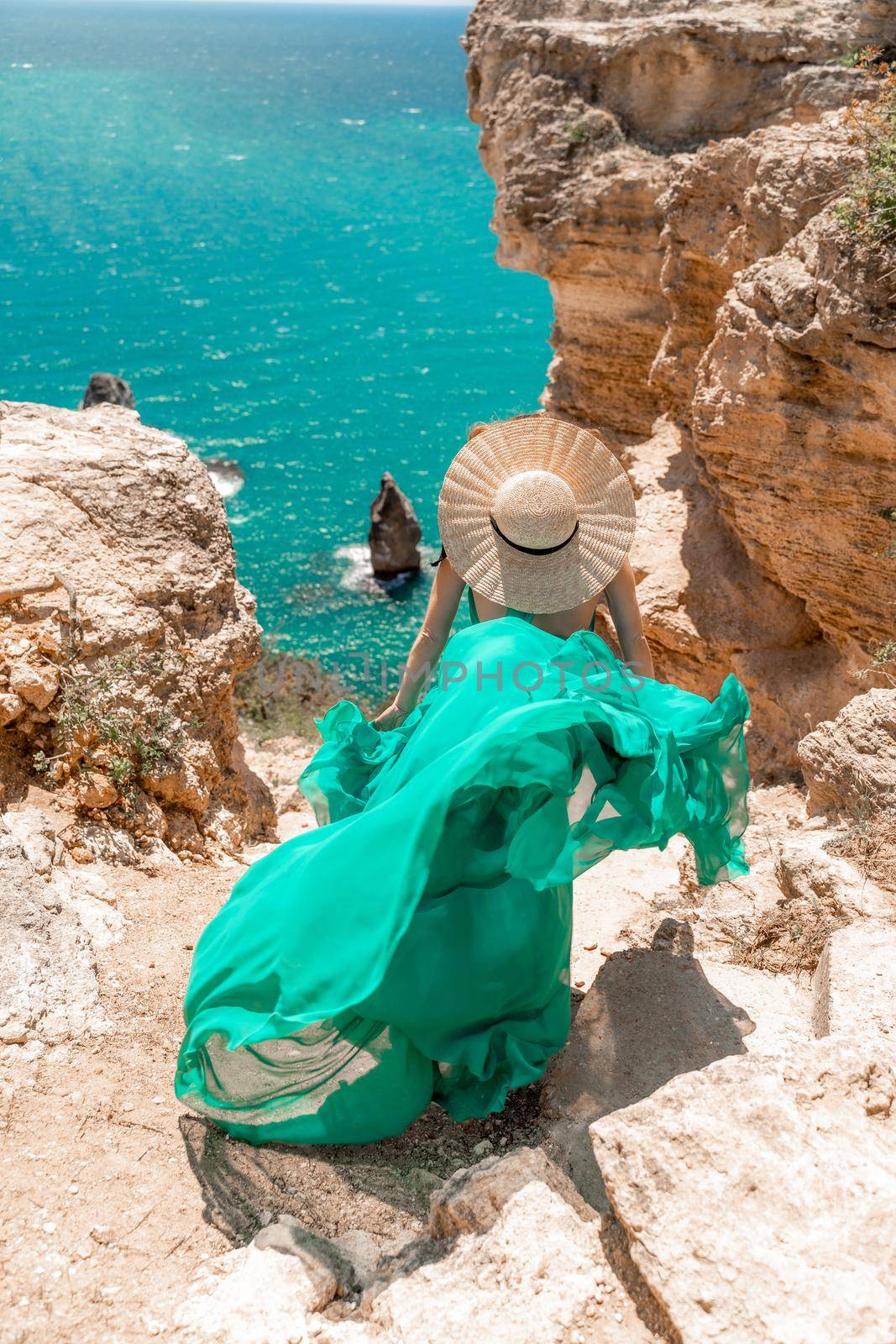 A girl with loose hair in a long mint dress and a straw hat descends the stairs between the yellow rocks overlooking the sea. A strong wind develops the dress. A rock can be seen in the sea