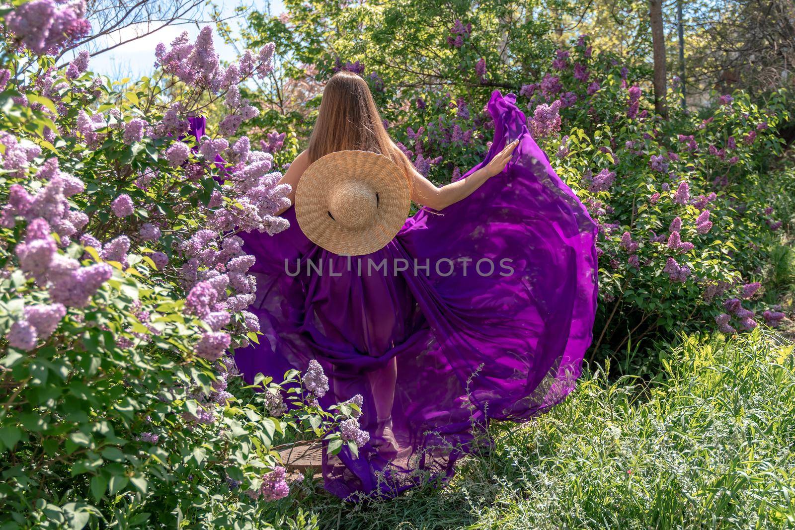 Fashion Model in Lilac Flowers, Young Woman in Beautiful Long Dress Waving on Wind, Outdoor Beauty Portrait in Blooming Garden by Matiunina