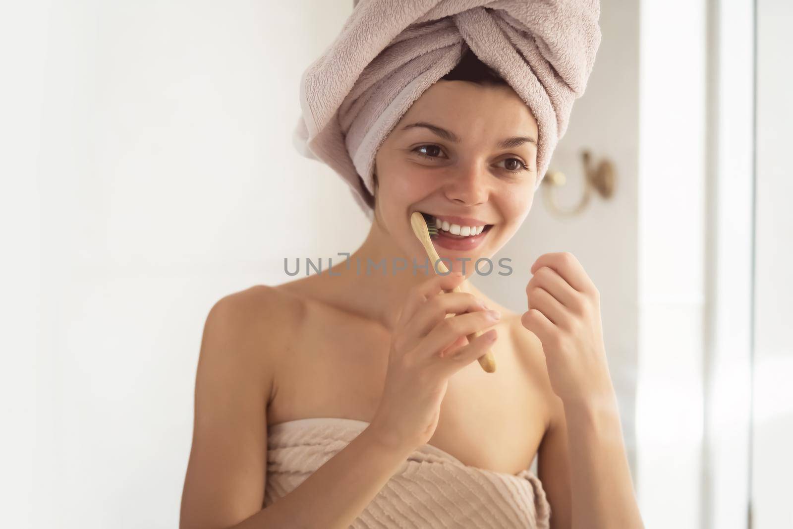 A young girl brushes her teeth near the mirror after relaxing in the bathroom, the woman is wrapped in a towel and takes care of the health and beauty of her body and teeth, gums.