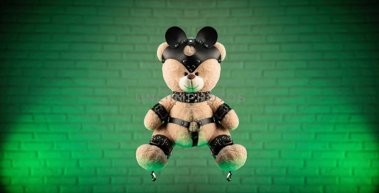 the toy Teddy bear dressed in leather belts and mask accessory for BDSM games on a light background texture of a brick wall