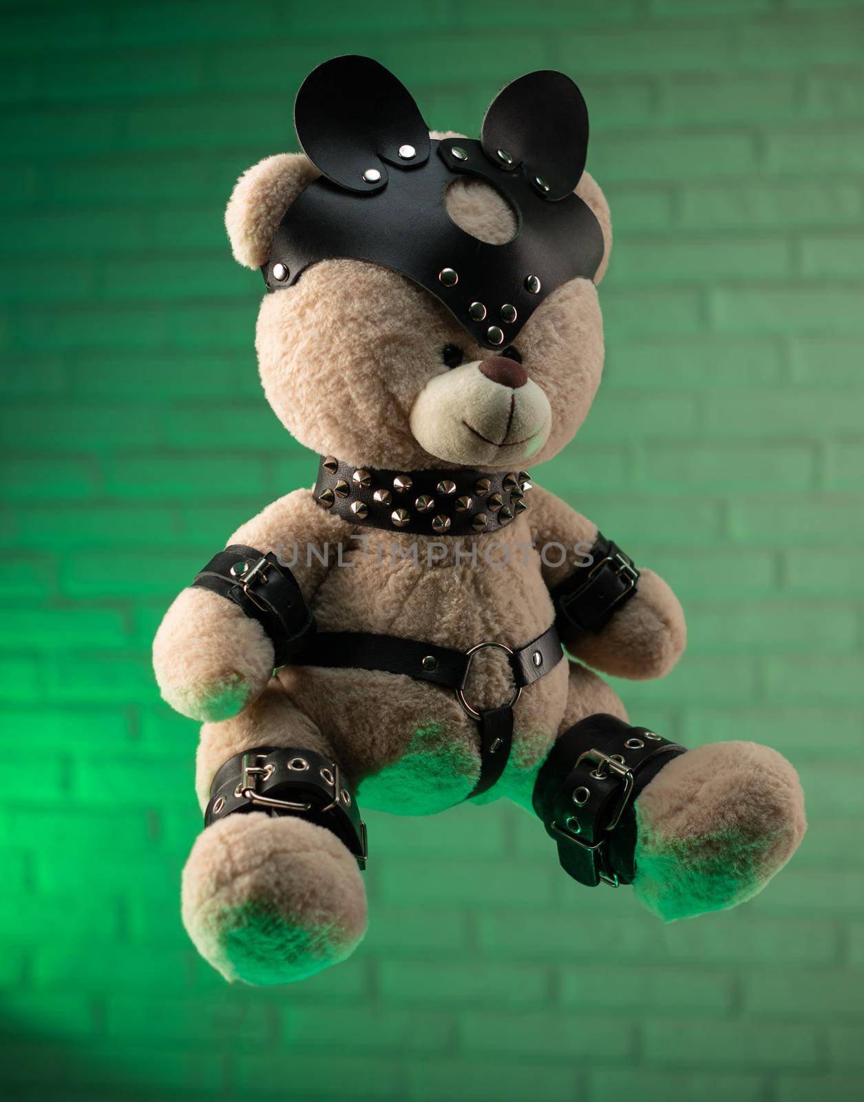 the toy Teddy bear dressed in leather belts and mask accessory for BDSM games on a light background texture of a brick wall