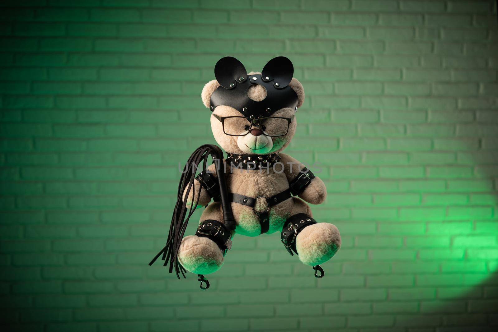 the toy Teddy bear dressed in leather belts and mask accessory for BDSM games on a light background texture of a brick wall