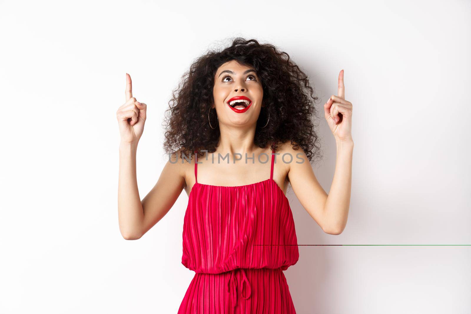 Excited caucasian woman with curly hair, red lips, looking and pointing up with amazed smile, checking out promo offer, white background.