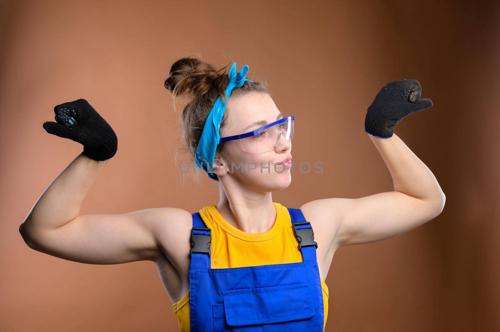 Strong stylish caucasian woman repairman foreman in overalls showing a gesture of strength showing biceps. Studio portrait of a young and strong feminine woman by yanik88