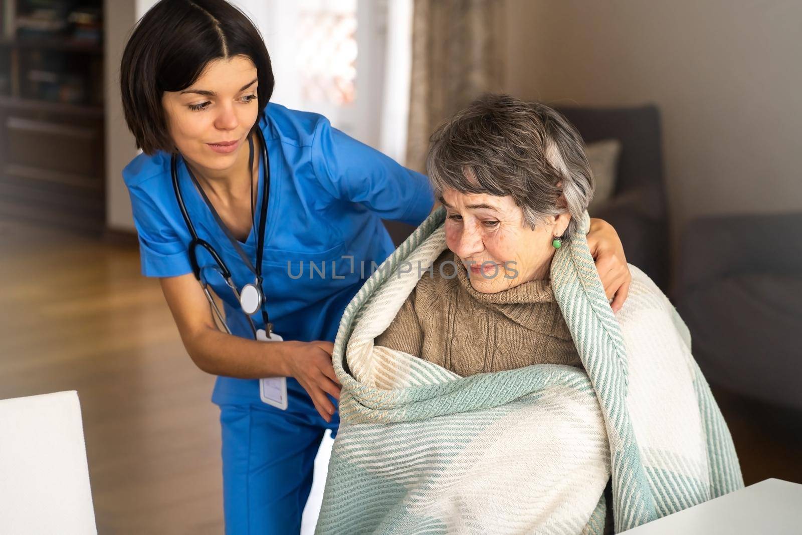 A young nurse takes care of an elderly 80-year-old woman at home, wraps a blanket around her. Happy retired woman and trust between doctor and patient. Medicine and healthcare.