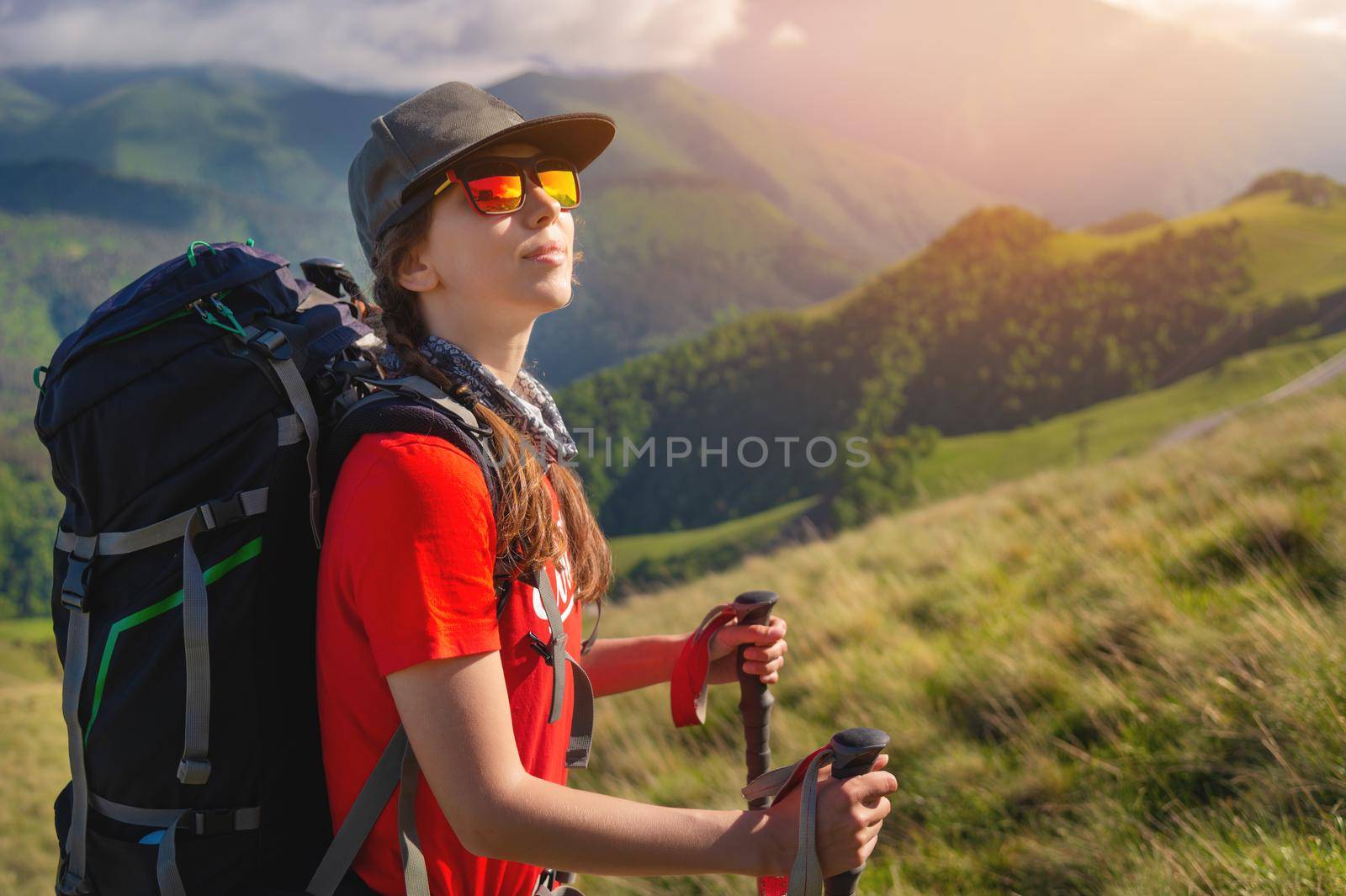 A tourist girl with a backpack admires the sunset from the mountainside. Traveler in the background of mountains and clouds at sunset. Woman tourist in sunglasses, cap and with a backpack stands on the slope of the mountain. Trekking and hiking in the mountains