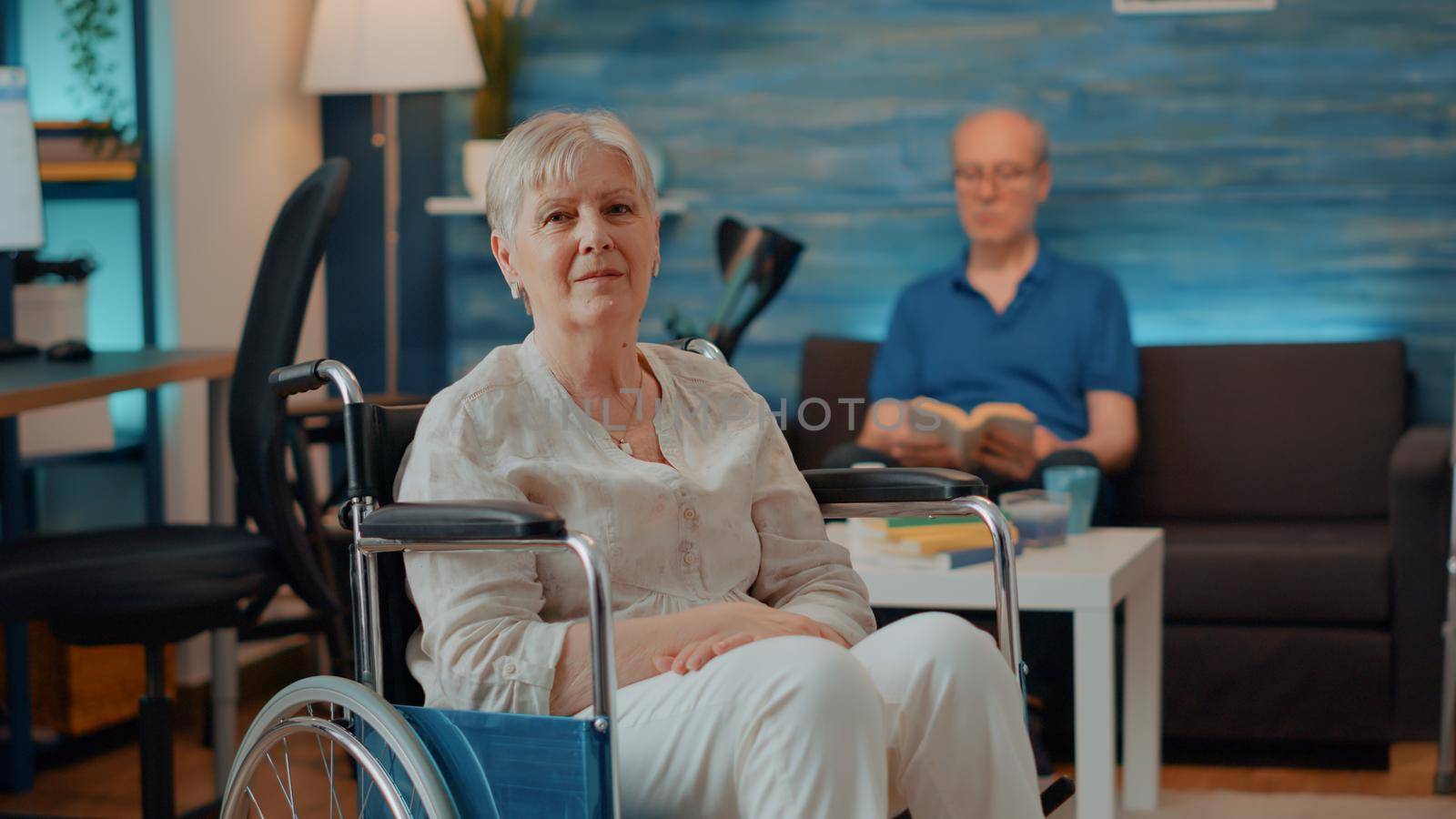 Portrait of older person with chronic disability looking at camera in living room. Senior woman in wheelchair suffering from mobility problem, using object to give support with transportation.