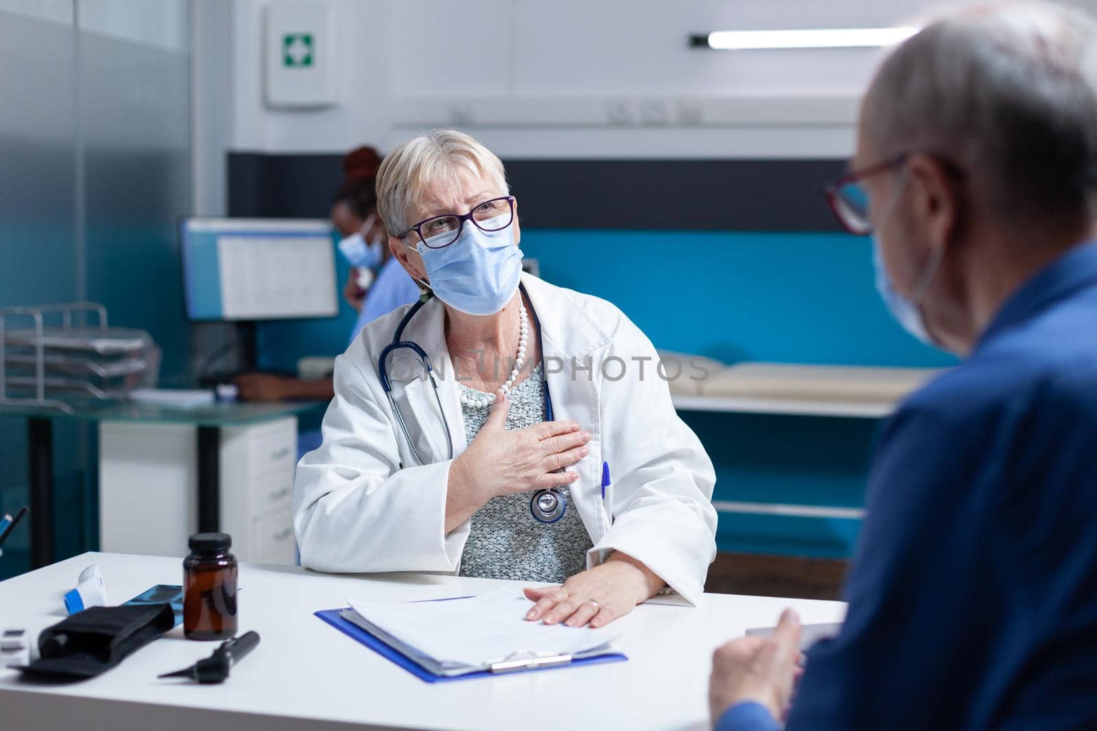 Woman doctor having checkup appointment with old patient, wearing face mask. Physician having conversation with man about healthcare and medical diagnosis in cabinet during pandemic.
