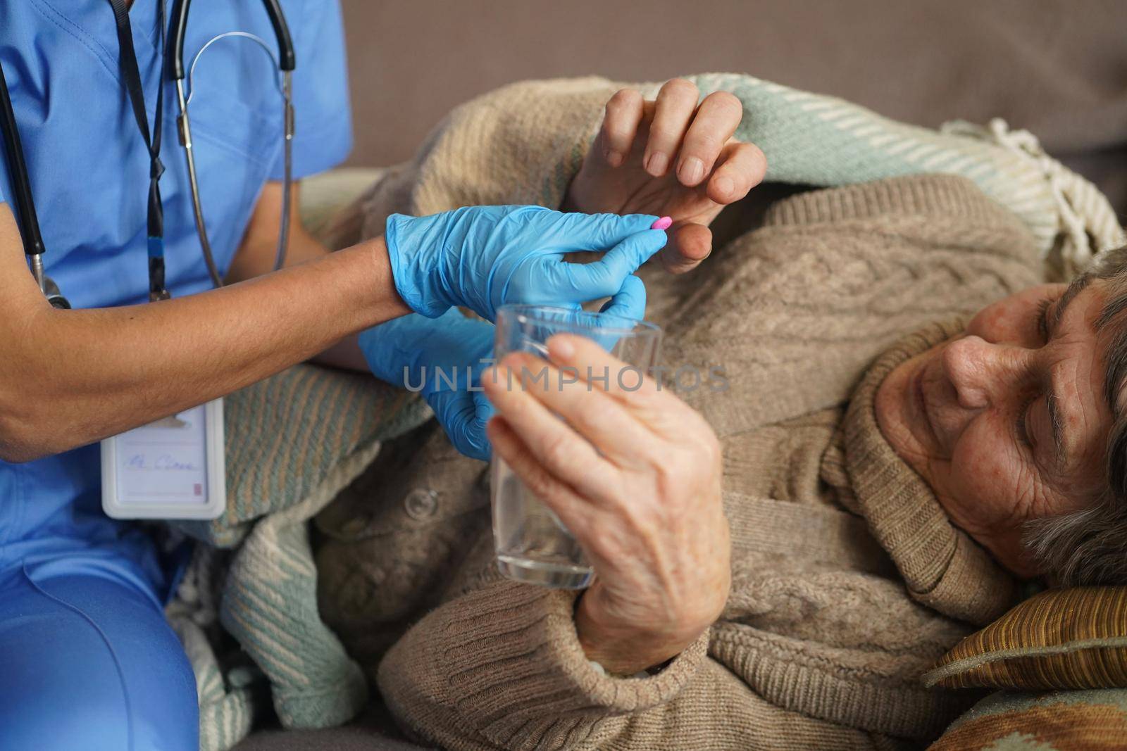 A young nurse is caring for an elderly 80-year-old woman at home. She holds a glass of water and gives medicine pills a pensioner retired woman who lies and rests in bed.