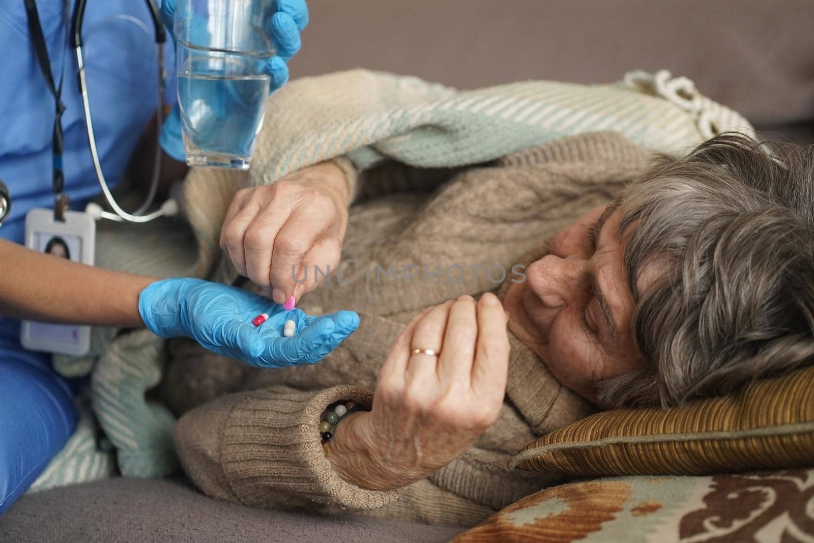 A young nurse is caring for an elderly 80-year-old woman at home. She holds a glass of water and gives medicine pills a pensioner retired woman who lies and rests in bed.