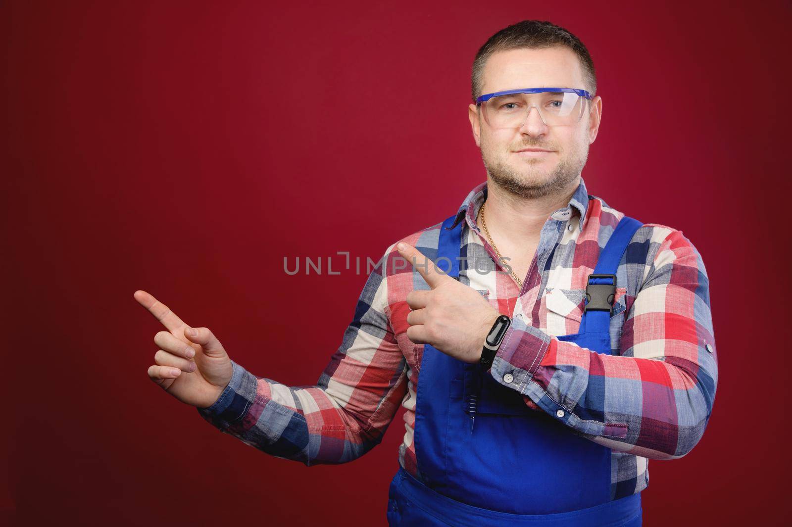 Attractive Caucasian male foreman in uniform and goggles points fingers at blank space. Foreman repairman maste studio portrait by yanik88
