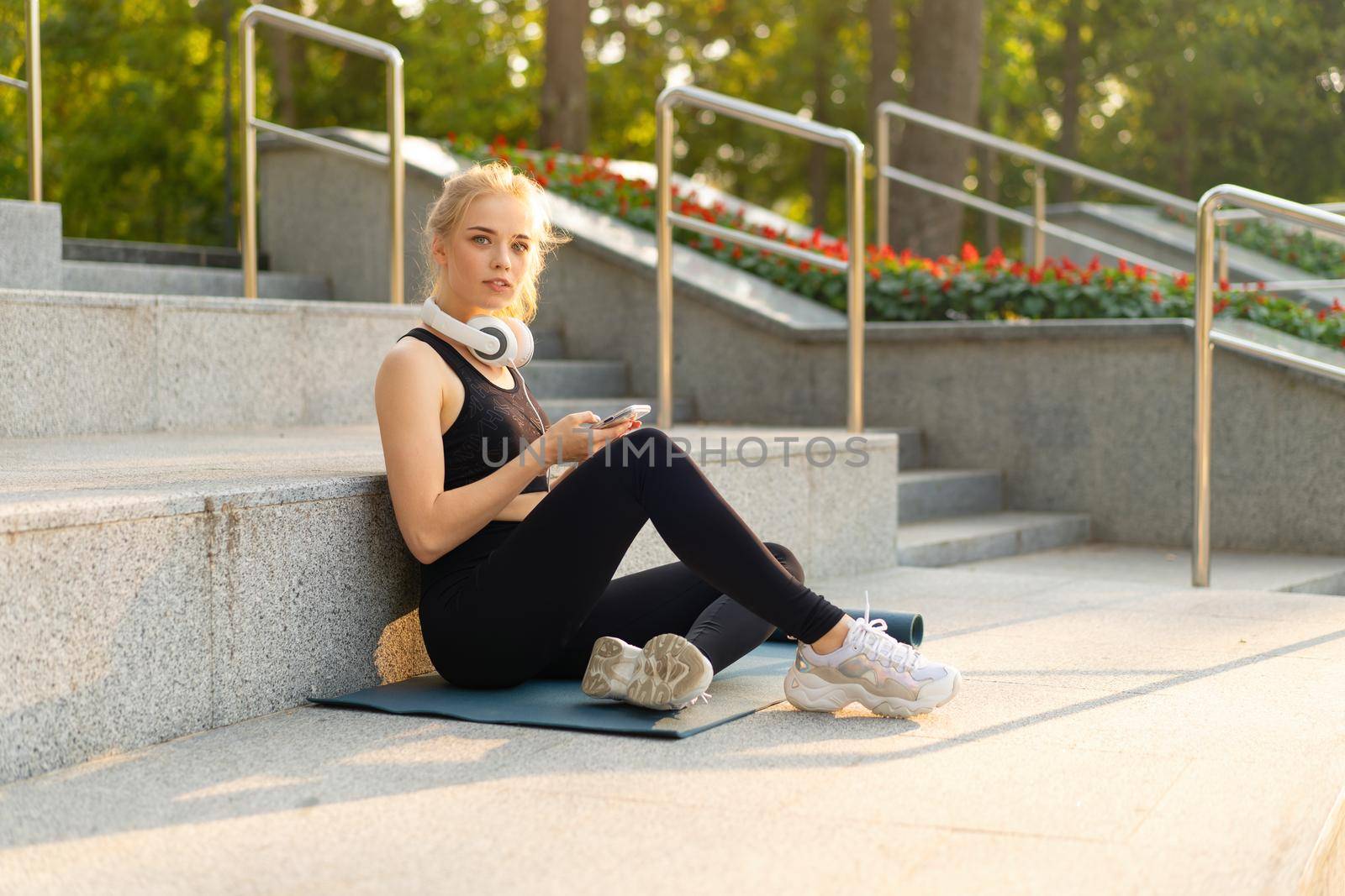 Sport and Fitness Young Adult Caucasian Woman Using Smartphone Sitting Exercise Mat Concrete Floor Resting after morning Workout Outdoor Summer Park Athletic Female Take a Break Use mobile Phone