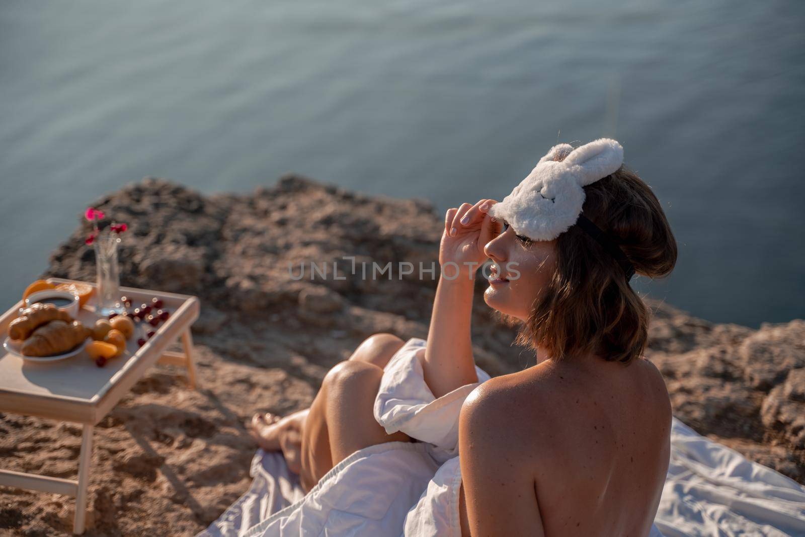 Young woman in sleep mask posing on the seashore. She is wrapped in a white blanket. The coast of the sea at a height, around the sea and rocks, dawn