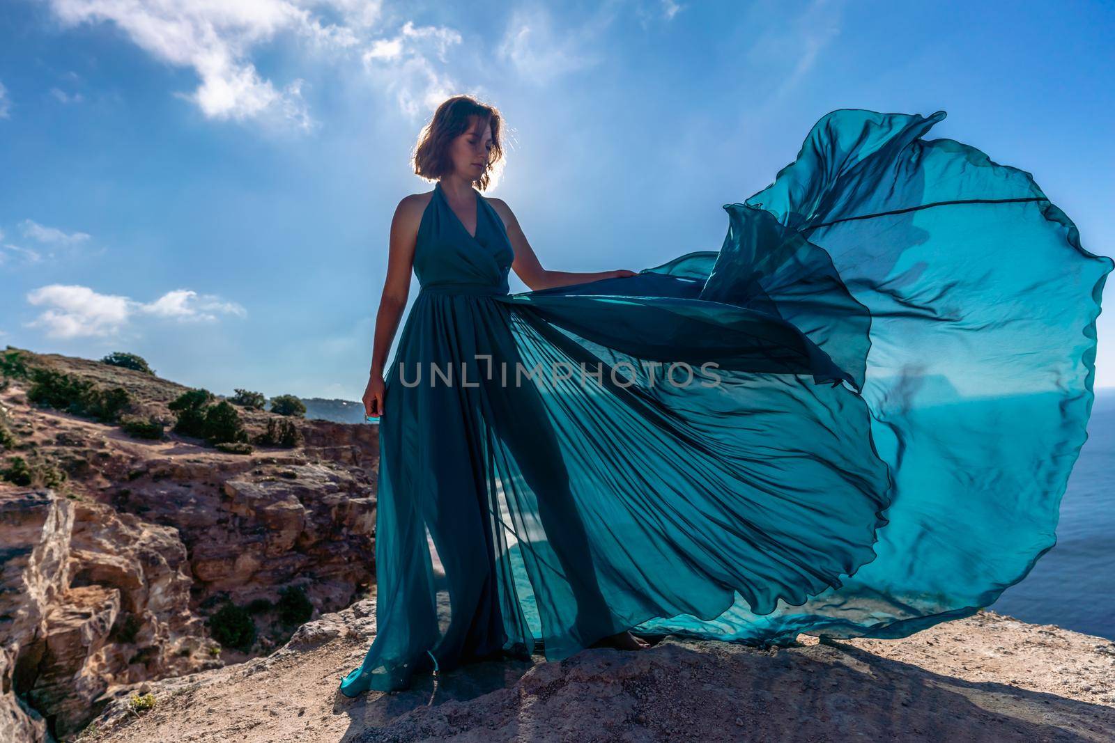 A girl with loose hair in a long mint dress descends the stairs between the yellow rocks overlooking the sea. A rock can be seen in the sea. Sunny path on the sea from the rising sun.