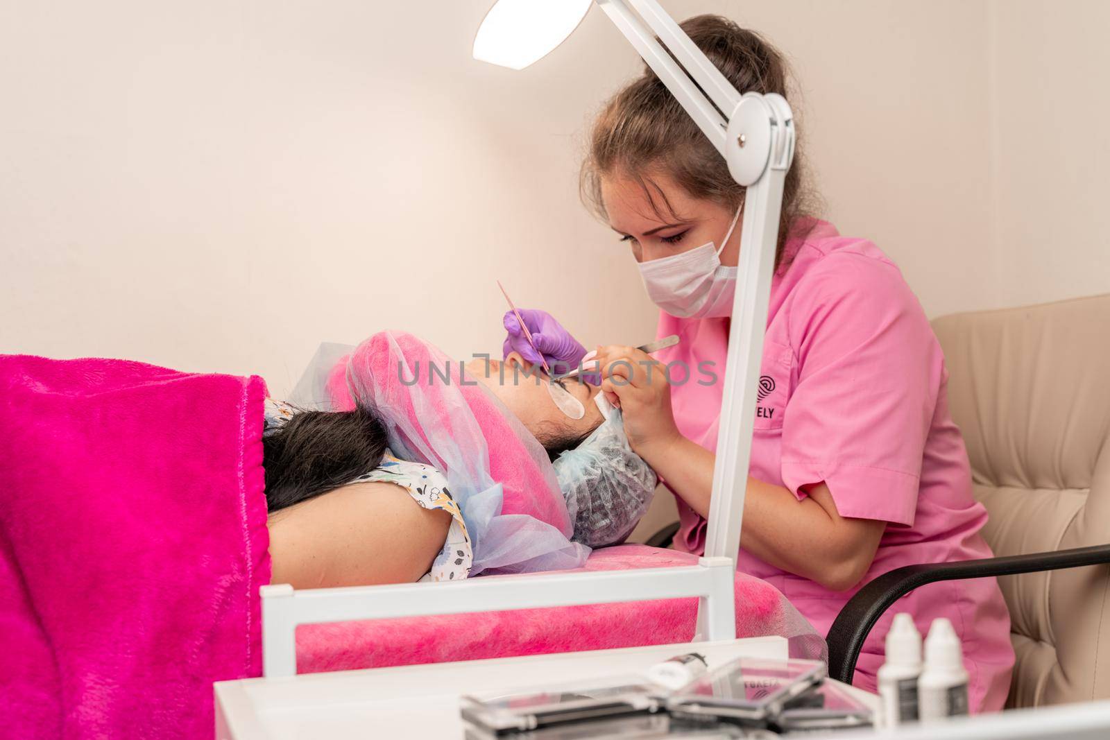 Master lashmaker, diligently extends eyelashes to the client, on the couch, in the beauty salon. A beauty master, a girl of 30 years old, European appearance, at work, in a bright room.