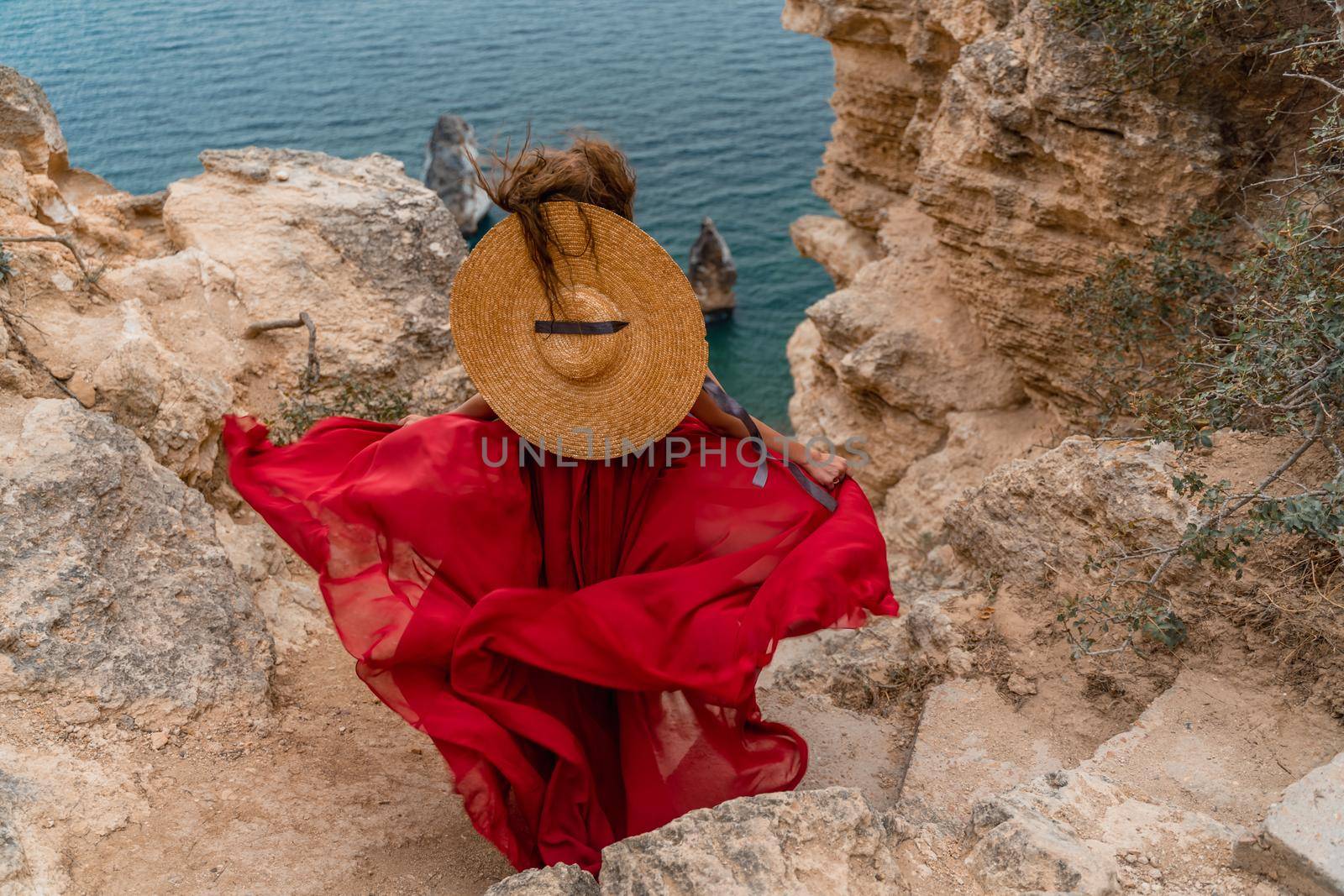 A girl with loose hair in a long red dress descends the stairs between the yellow rocks overlooking the sea. A rock can be seen in the sea. Sunny path on the sea from the rising sun.