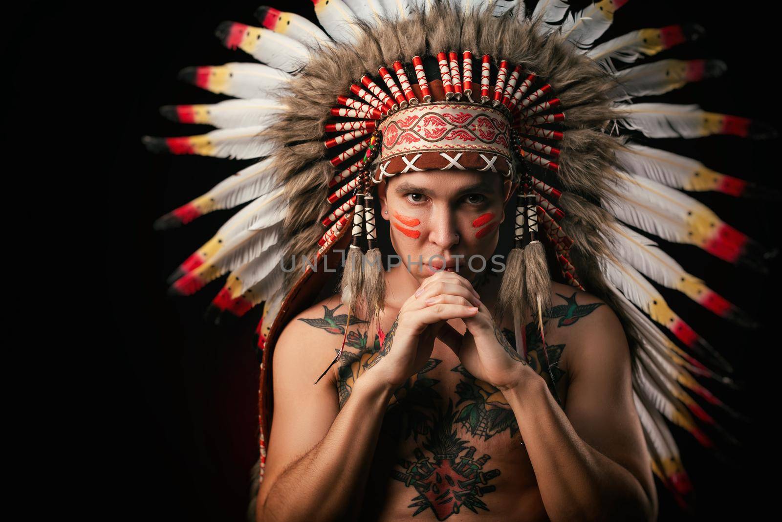 white man in a headdress of the indigenous peoples of North America with a naked torso on a dark background