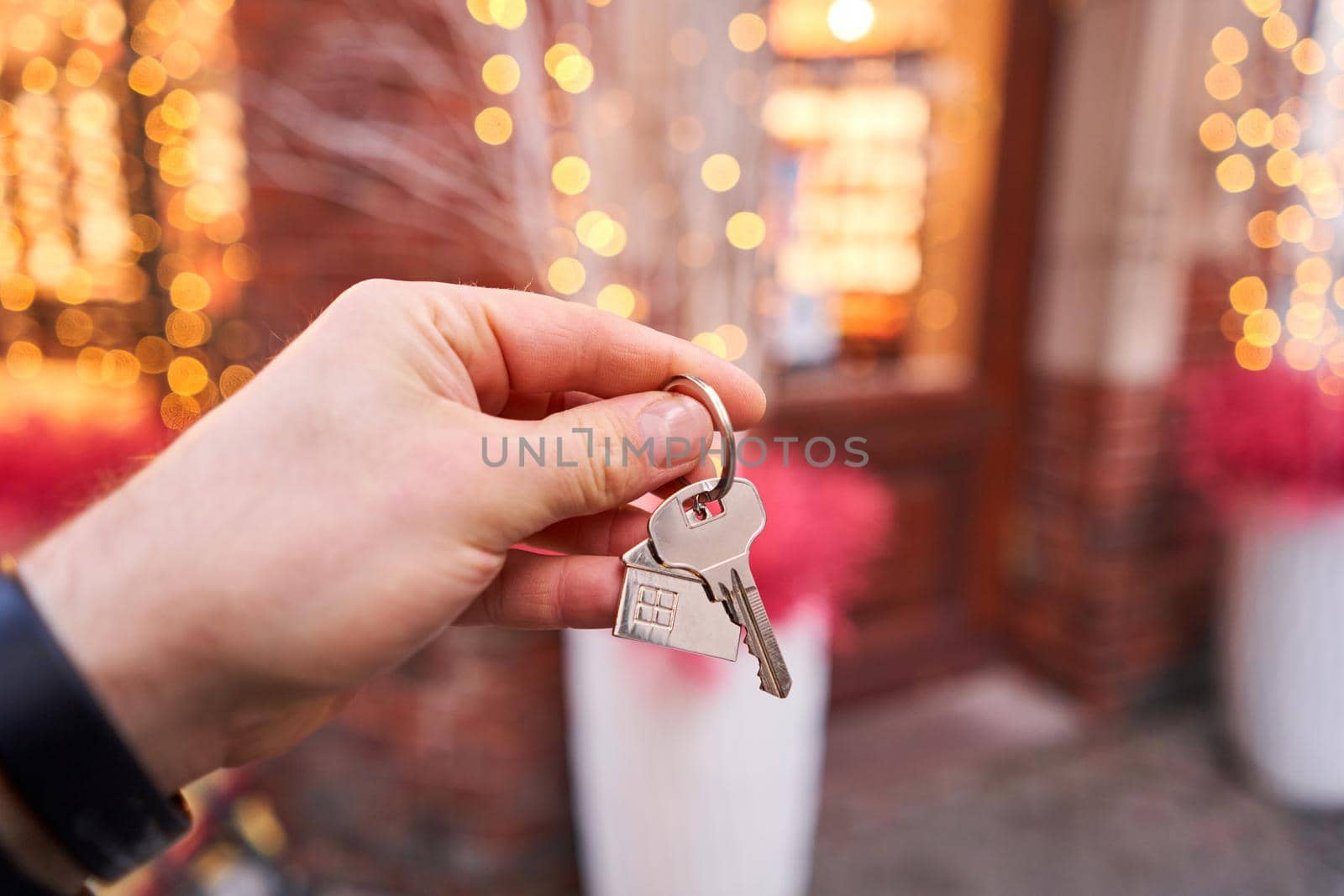 Rent a house for Christmas. Mortgage or rent concept. Man holding key with wooden house keychain . Real estate, hypothec, moving home or renting property. Christmas mood in blurred background. by Malkovkosta