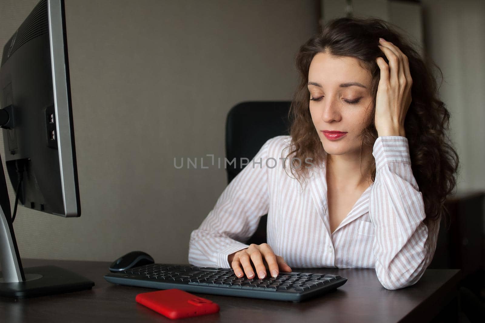 Young female office manager in white shirt and curly hair is sitting at the table and typing using keyboard, routine work, freelance