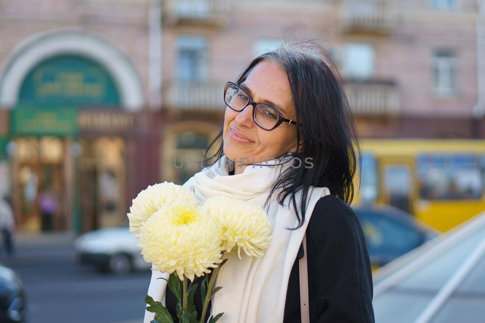 A beautiful middle-aged woman getting grey-haired in a dark coat in a spring town with a bouquet of flowers