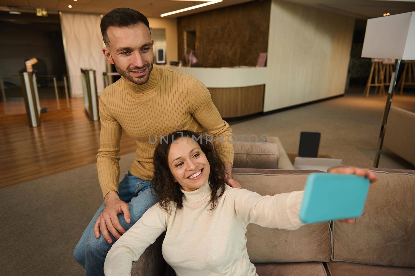 Attractive Hispanic woman sitting next to her boyfriend and making a self-portrait on smartphone while resting together in the lounge zone of the airport departure terminal while waiting for a flight