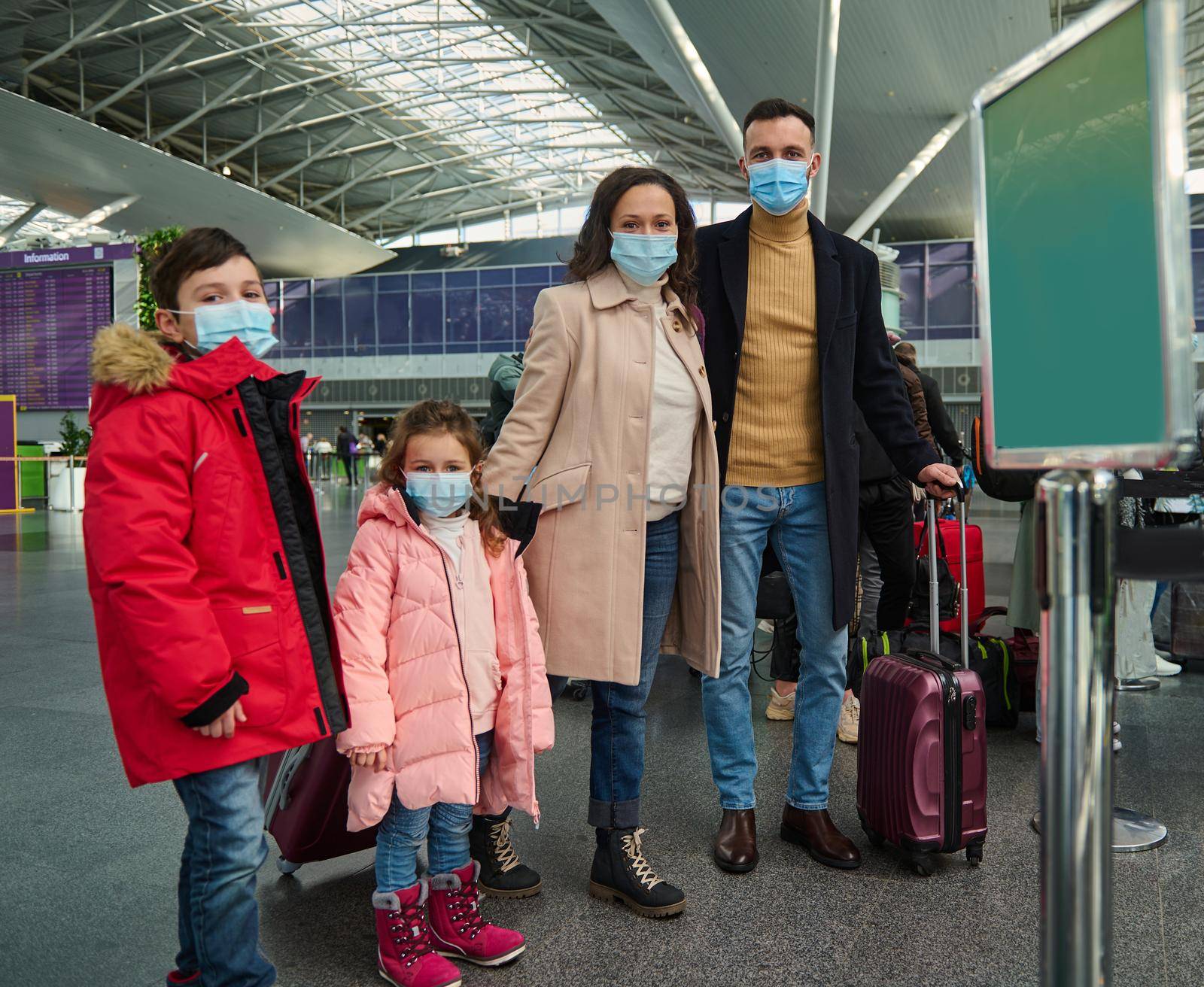 Happy Caucasian family of father, mother, son and daughter wearing protective medical mask dressed in warm clothes standing with luggage and suitcases at the airport and waiting check-in for flight