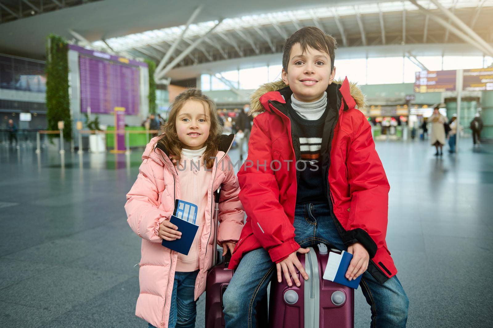 Cheerful Caucasian children- boy and girl in warm clothes, sitting on a suitcase and holding passports, with air tickets and boarding pass at the international airport. Family airplane travel concept by artgf