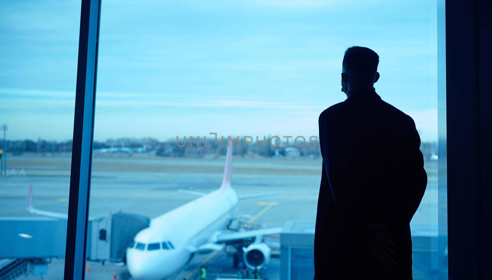 Silhouette portrait of a businessman during business trip standing by the panoramic window in the airport lounge, looking at the plane and the runways while waiting for his flight by artgf