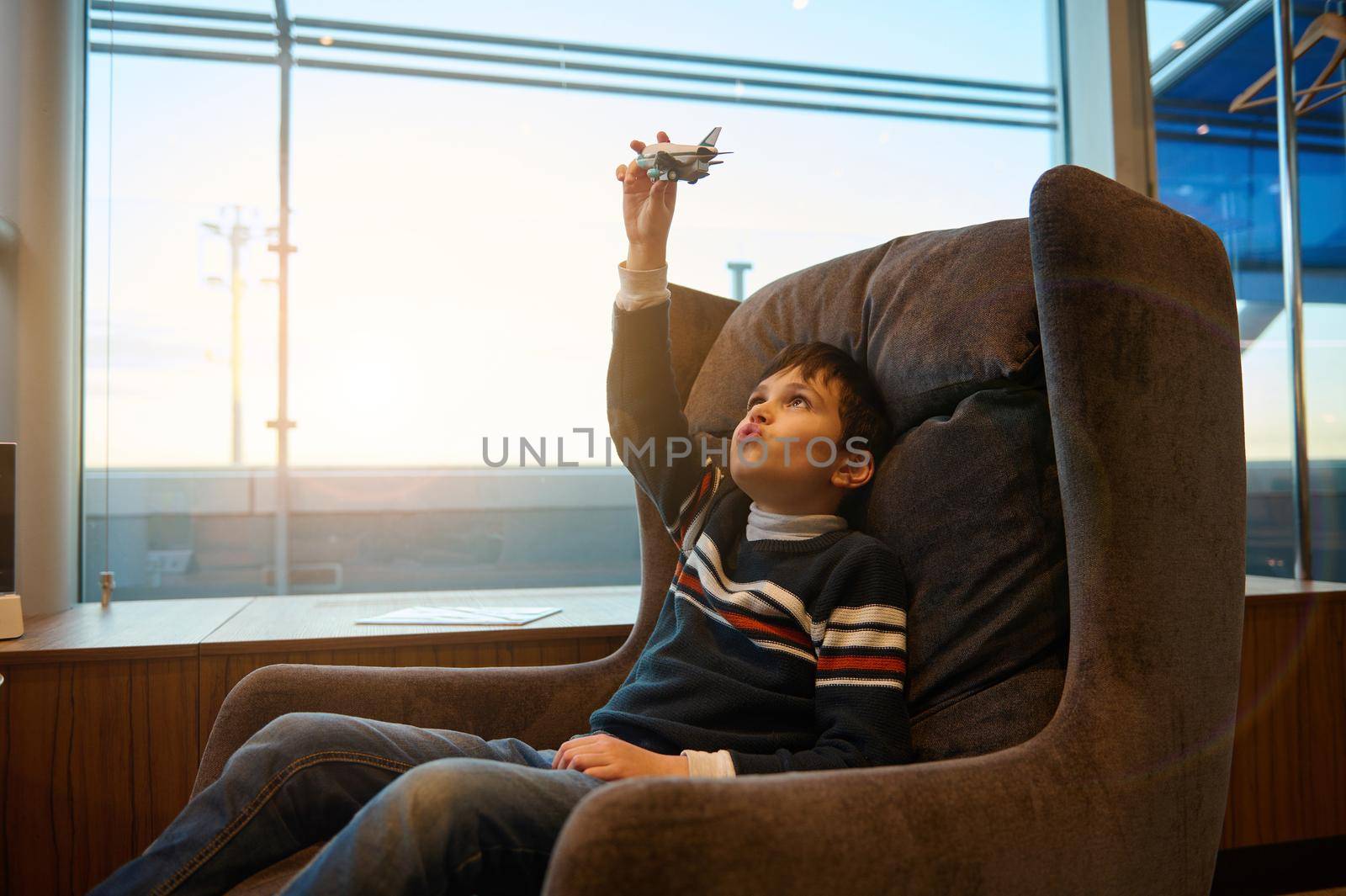 Handsome European teenage boy plays with toy airplane sitting in a chair by panoramic windows overlooking the runway at sunset while waiting to board flight at international airport departure terminal