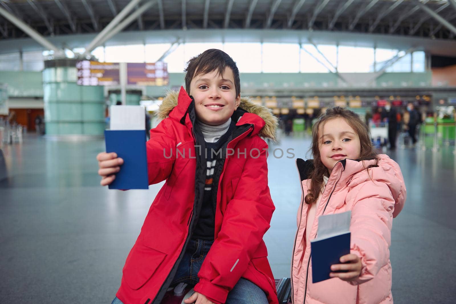 Handsome teenage boy and cute little girl wearing warm down jackets smiling toothy smile while standing at international airport and holding passport documents with air tickets and boarding pass