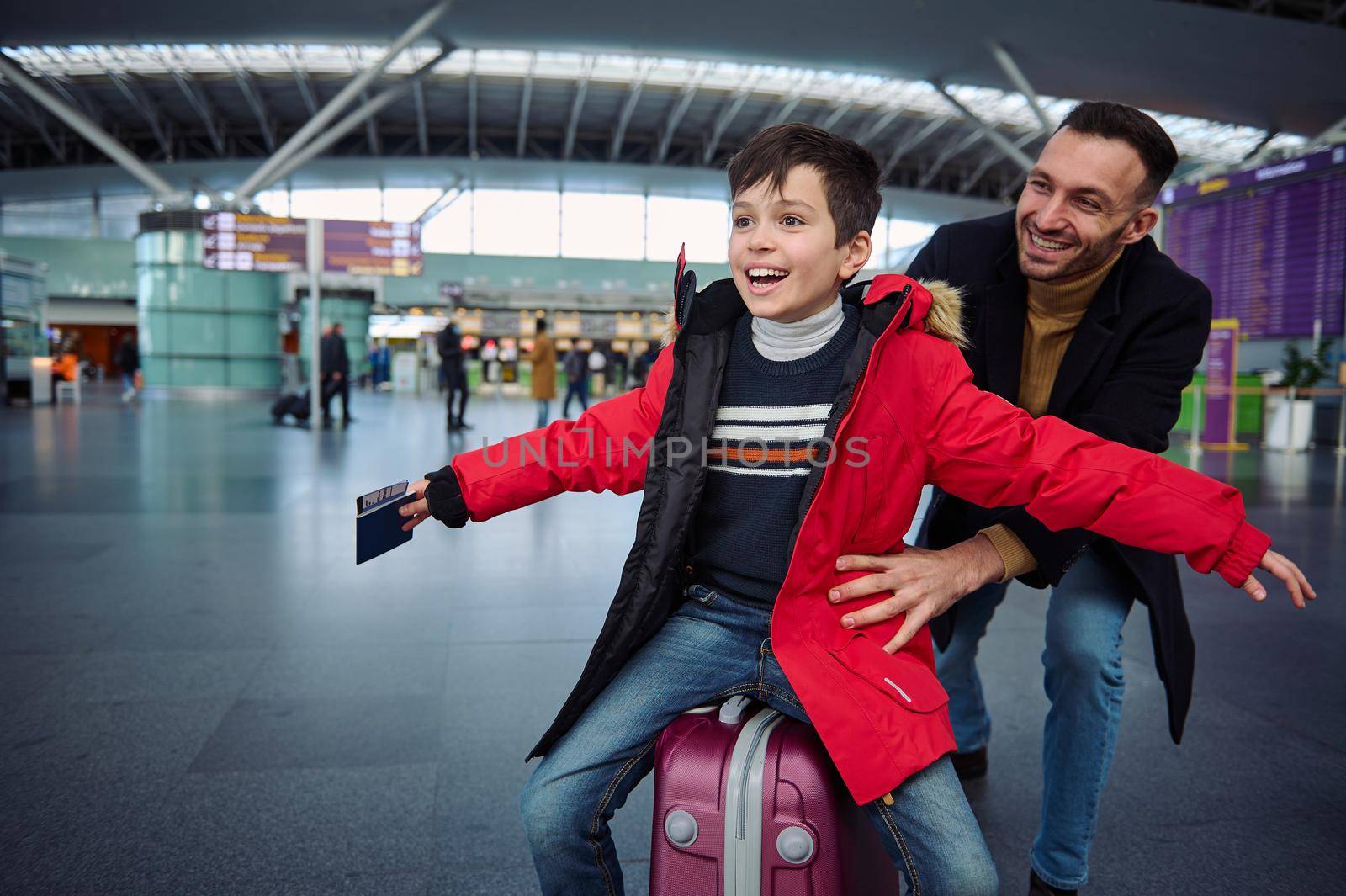 Charming little boy with a passport and a boarding pass in his outstretched hands while his dad is riding him on a suitcase in the departure hall of the airport, awaiting check-in for a flight