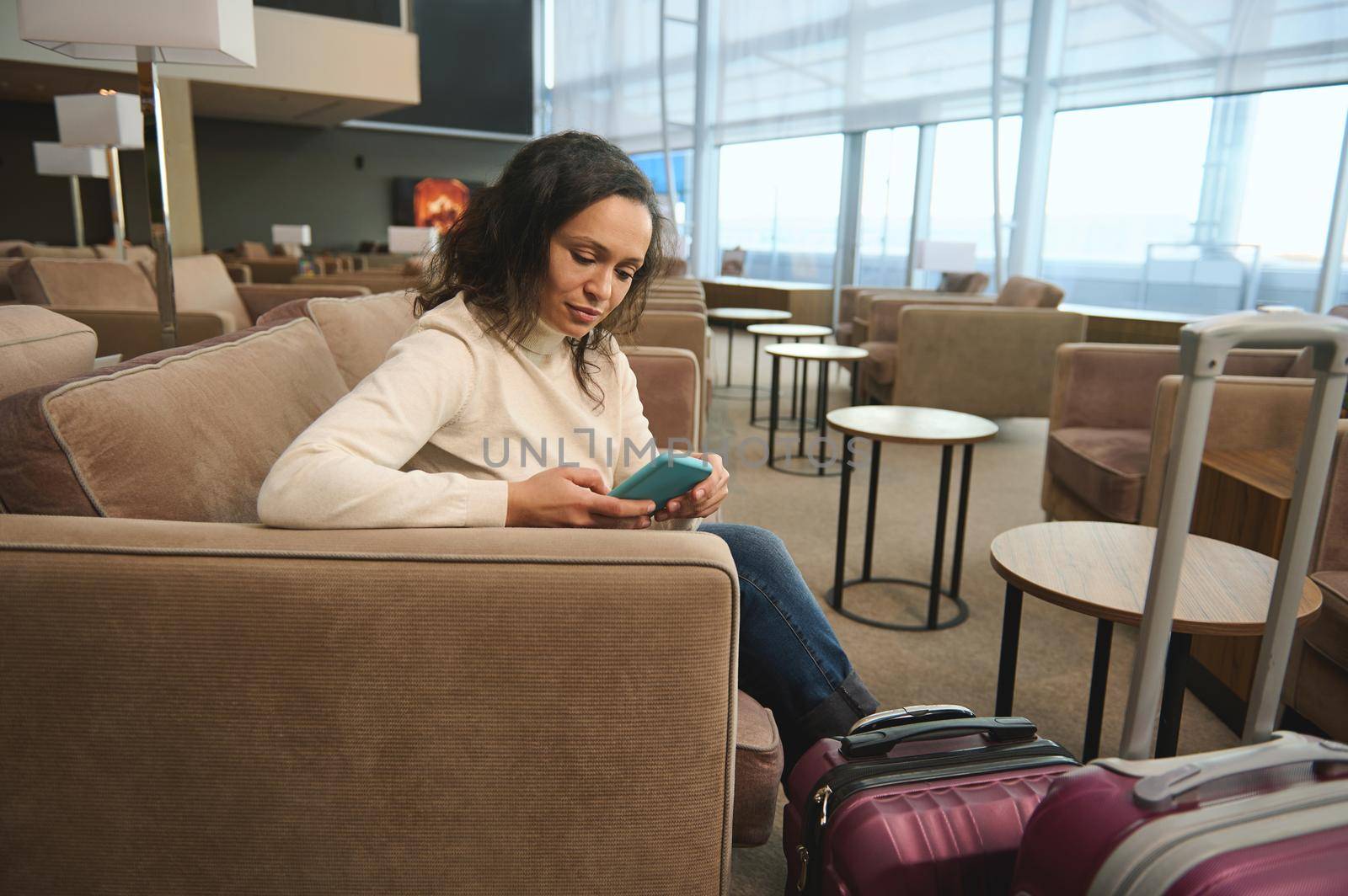 Confident business woman traveling alone, sitting at a table and using a mobile phone, relaxing on an armchair in the luxurious lounge of the airport departure terminal while waiting to board a flight by artgf