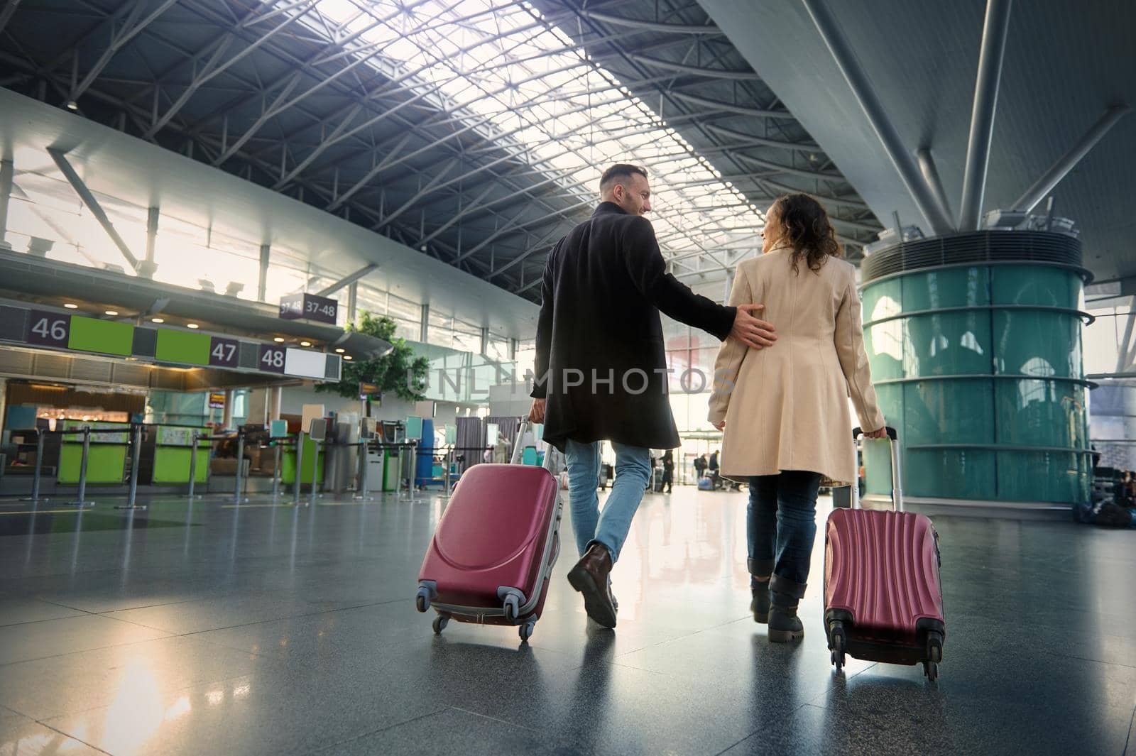 Full-length of two handsome people man and woman, young couple with suitcases walking along the departure hall of international airport on the blurred background of flight check-in counters