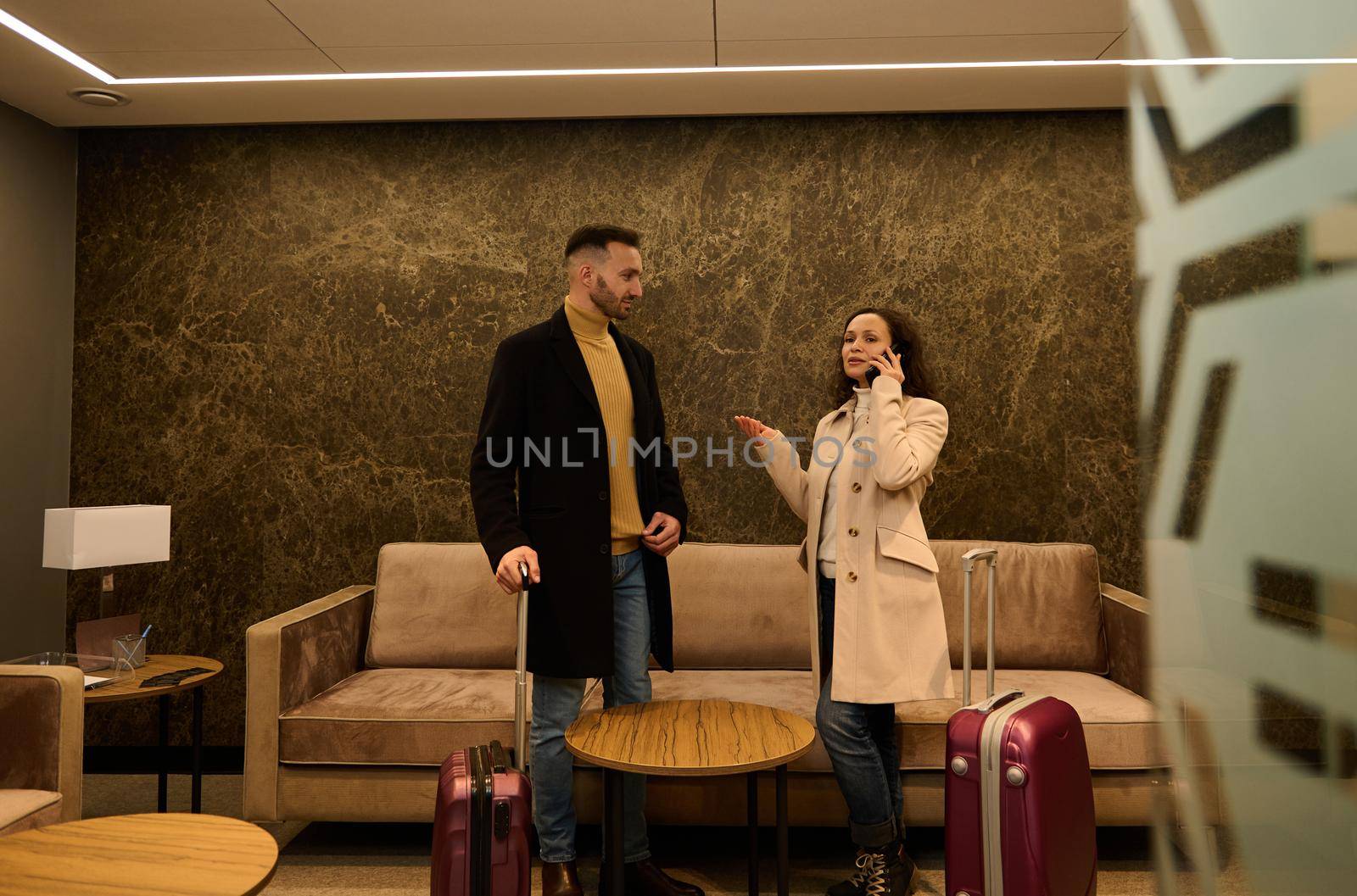 Delighted newlywed couple in the VIP lounge of the airport terminal. Handsome business man looks at his partner who is on the phone, negotiating and booking a hotel while waiting to board a flight