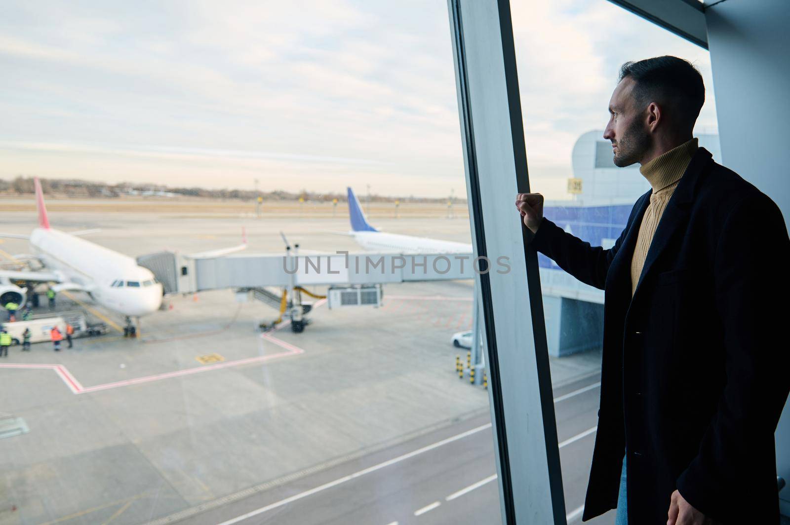 Confident handsome businessman on a business trip stands at the panoramic window in the waiting room of the international airport, admiring the planes on the runways while waiting to board the flight by artgf