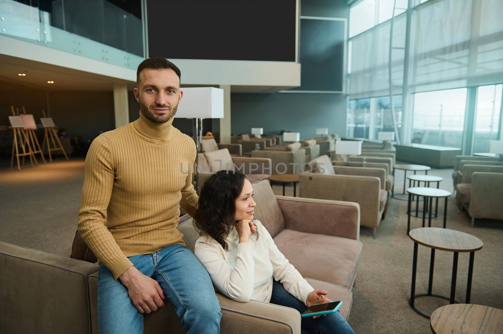 Attractive handsome Caucasian man looks at camera hugging his girlfriend, sitting in chair in an international airport lounge, waiting to board a flight. Multi ethnic couple travels on their honeymoon
