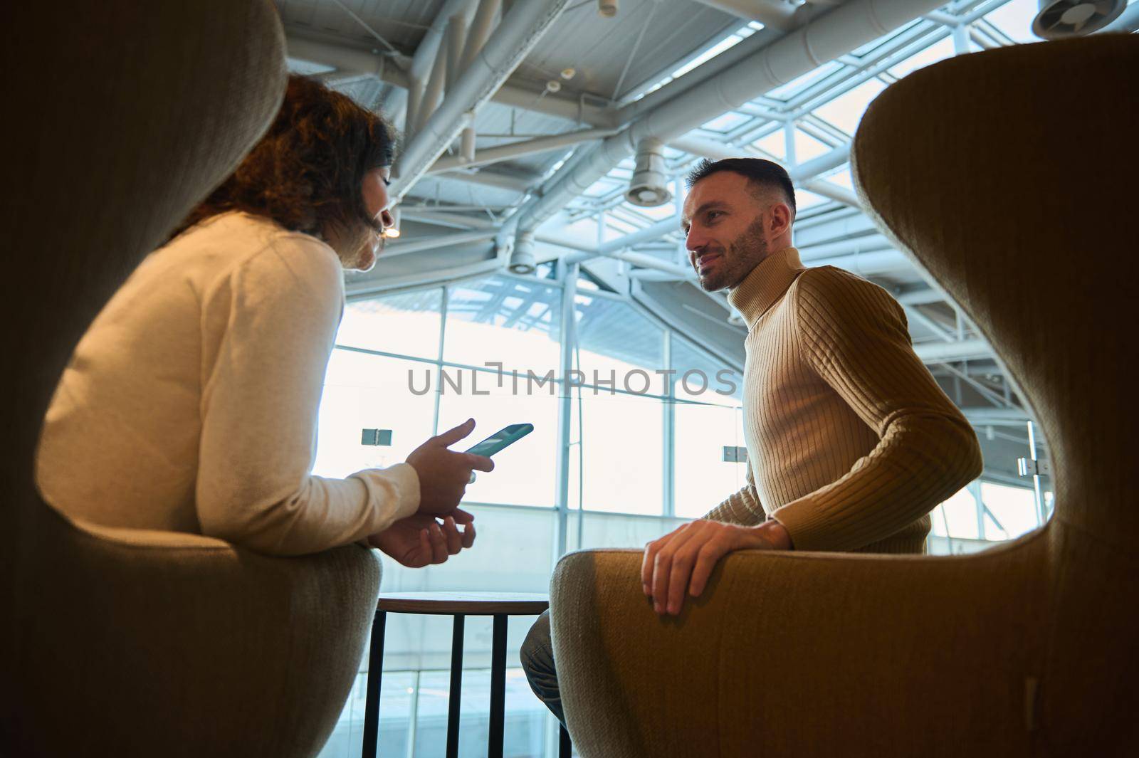 Happy couple in love, business partners, handsome man and pretty woman resting together, sitting on armchairs in airport departure terminal lounge talking together while waiting to board the flight.