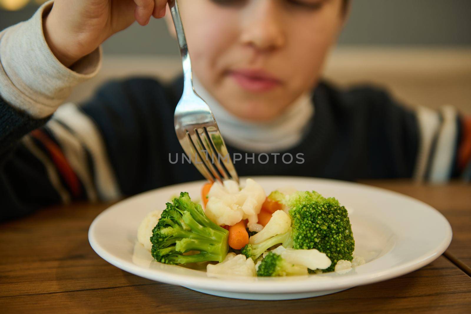 Nutrition, vegetarian diet for toddler. Focus on a plate of steamed vegetables, healthy meal of broccoli and carrots against blurred boy eating sitting at a table and holding fork